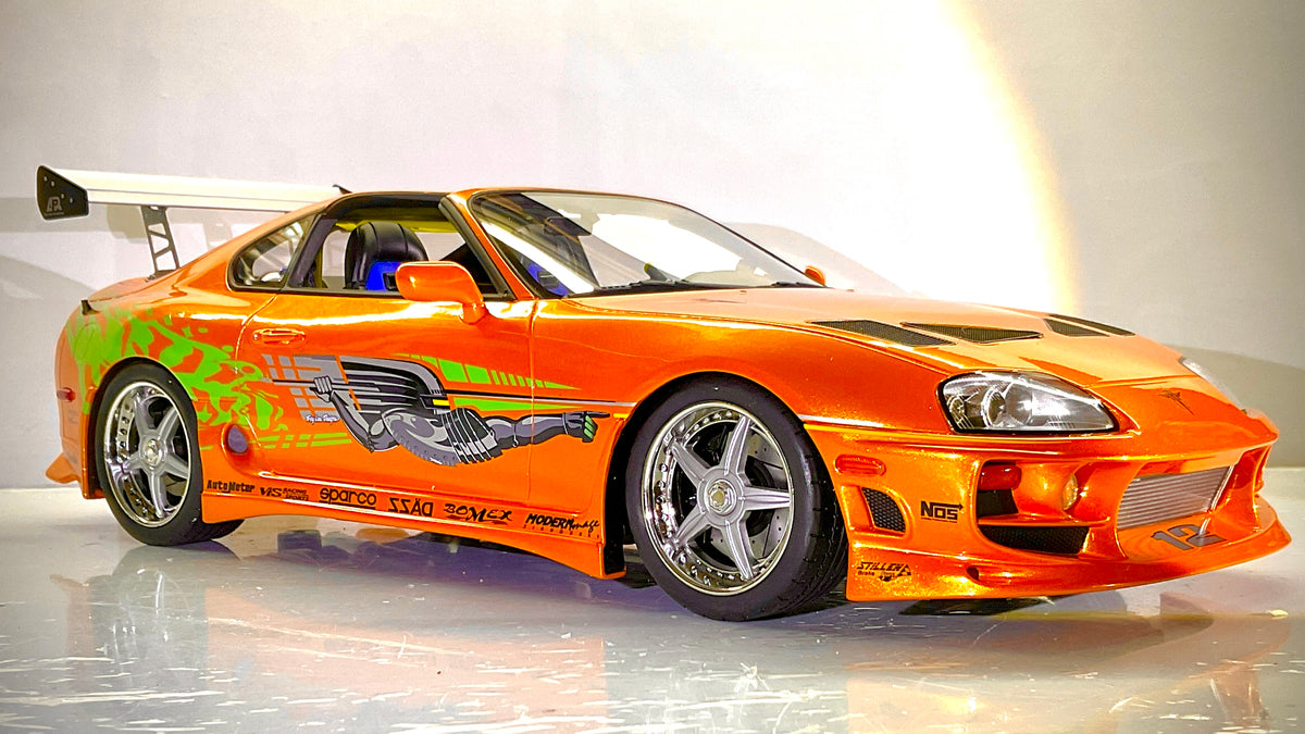 Foto a tre quarti senza scatola: Toyota Supra Fast & Furious arancione Kyosho 1:12, vista a tre quarti anteriore con grafiche laterali e alettone posteriore.